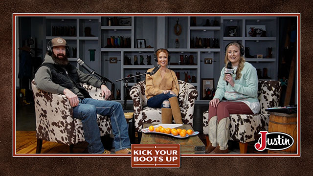 Two women and a man with a beard talking to each other, sitting down in cow print chairs near a coffee table, smiling at the camera.